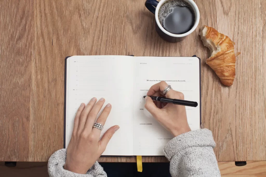 person-handling-pen-writing-notebook-beside-coffee-bread