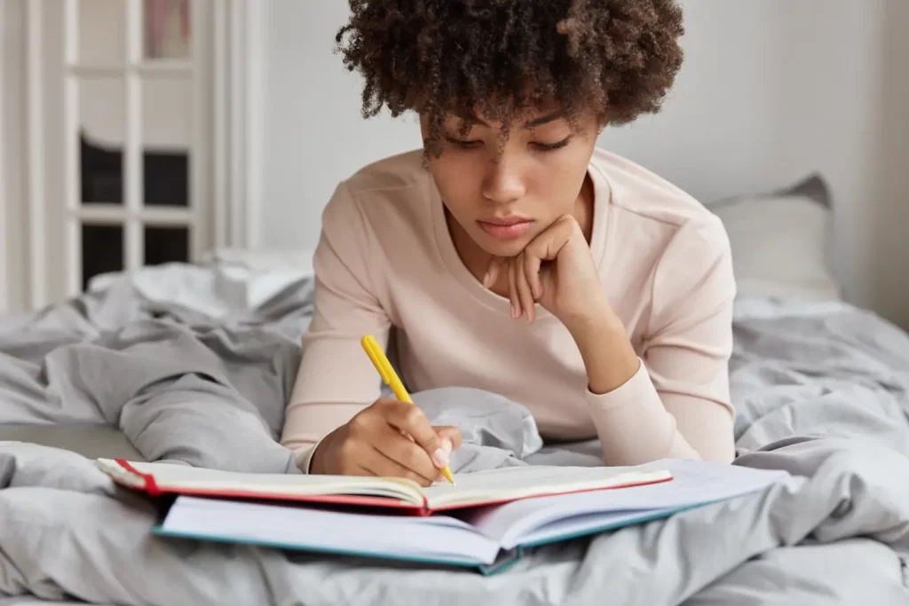 serious-dark-skinned-student-prepares-exam-from-early-morning-writes-important-notes-from-book-into-notebook-lies-unmade-bed-own-room