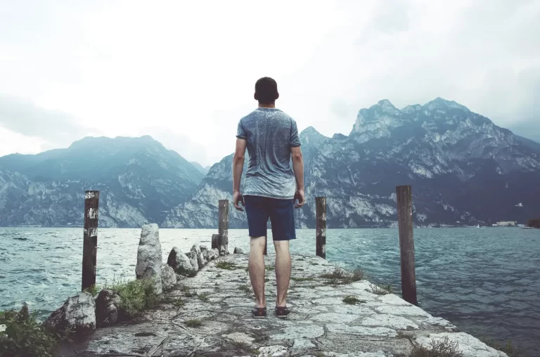 man-standing-on-gray-concrete-dock-facing-body-of-water-and-mountains-at-daytime